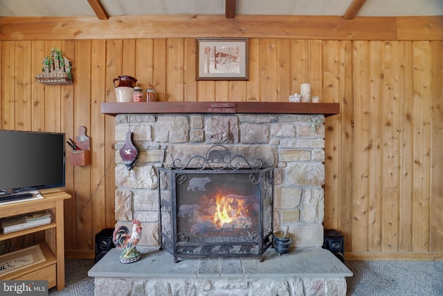 room details with beamed ceiling, dark colored carpet, wooden walls, and a stone fireplace