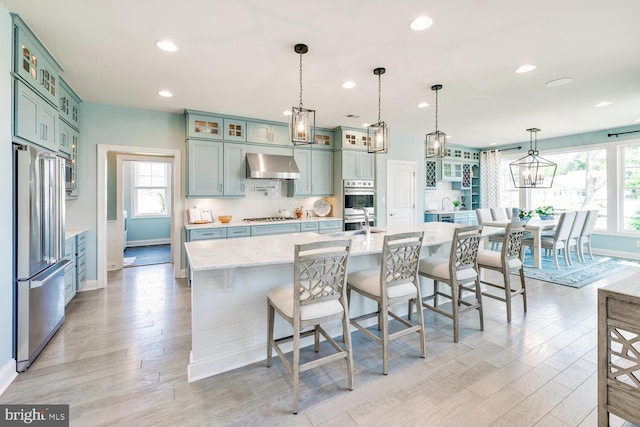 kitchen with hanging light fixtures, light hardwood / wood-style floors, stainless steel appliances, wall chimney exhaust hood, and a kitchen bar