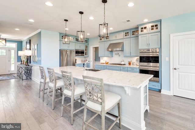 kitchen with decorative light fixtures, a kitchen island with sink, appliances with stainless steel finishes, a breakfast bar, and wall chimney range hood