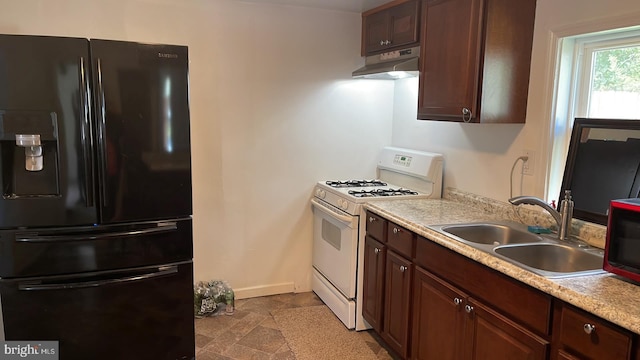 kitchen featuring white gas stove, sink, light tile floors, wall chimney range hood, and black fridge