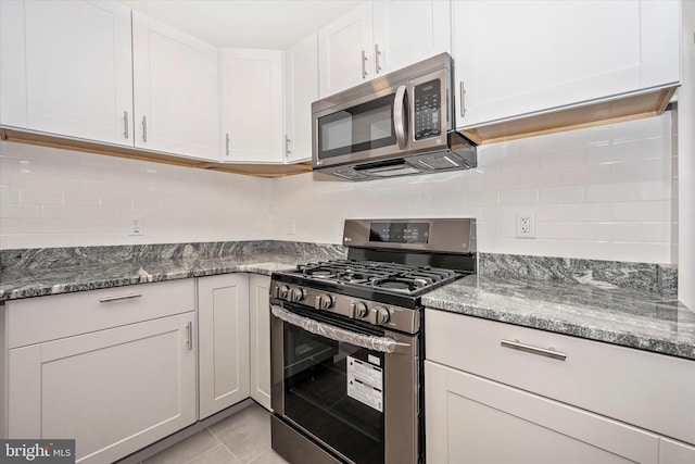 kitchen featuring dark stone countertops, stainless steel appliances, white cabinets, backsplash, and light tile floors