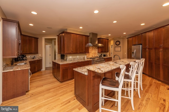 kitchen featuring wall chimney exhaust hood, light stone countertops, appliances with stainless steel finishes, light hardwood / wood-style floors, and an island with sink