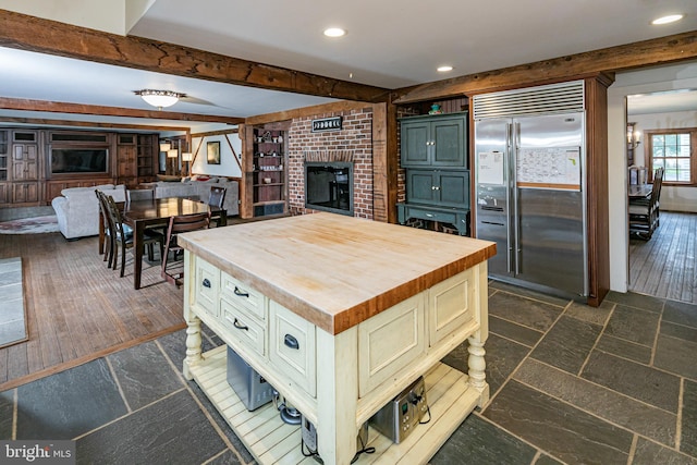 kitchen featuring a brick fireplace, a kitchen island, stainless steel built in fridge, wooden counters, and dark tile floors