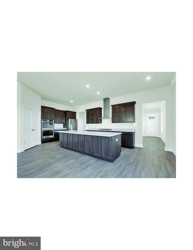 kitchen with dark brown cabinets, a kitchen island, appliances with stainless steel finishes, light hardwood / wood-style floors, and wall chimney range hood