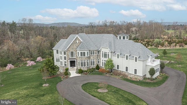 view of front of home featuring curved driveway, stucco siding, a balcony, stone siding, and a front lawn