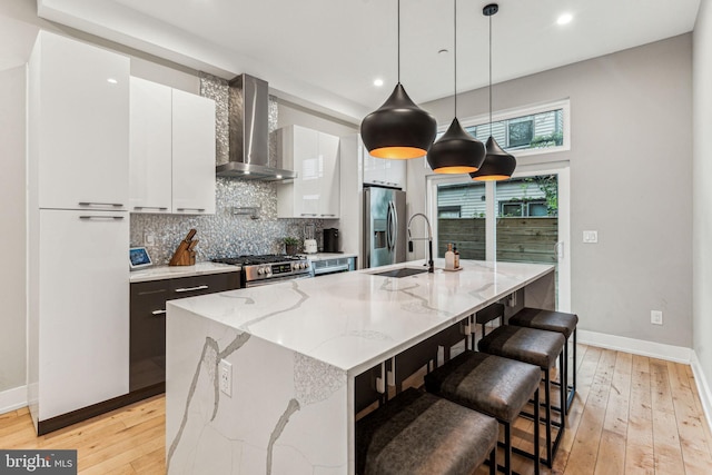 kitchen featuring wall chimney exhaust hood, white cabinetry, stainless steel appliances, a center island with sink, and light hardwood / wood-style floors