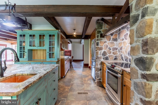 kitchen with a center island with sink, appliances with stainless steel finishes, beamed ceiling, light stone countertops, and sink