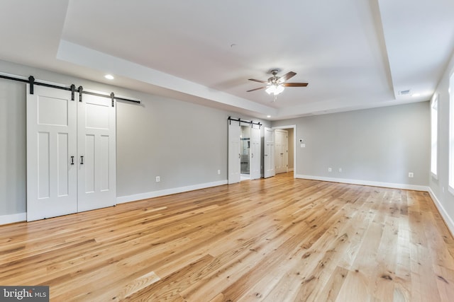 unfurnished bedroom featuring ceiling fan, a barn door, a raised ceiling, and light wood-type flooring