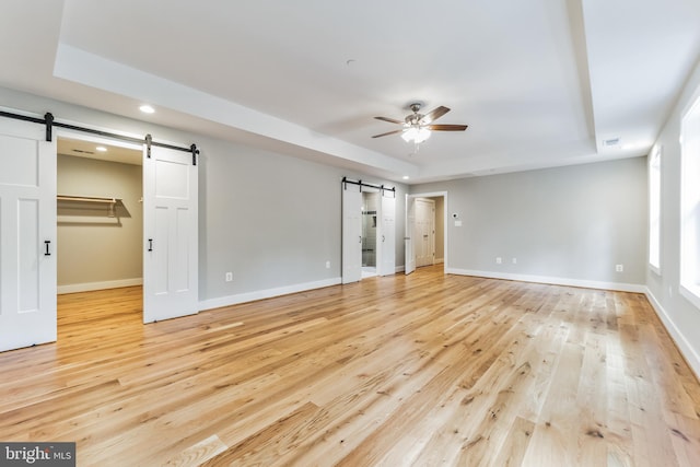 unfurnished bedroom featuring a raised ceiling, a barn door, light wood-type flooring, and ceiling fan