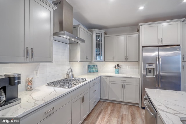 kitchen with appliances with stainless steel finishes, wall chimney exhaust hood, backsplash, light hardwood / wood-style flooring, and light stone countertops