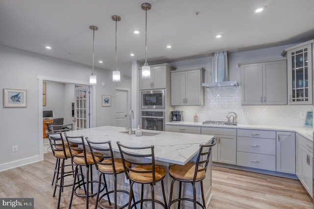 kitchen featuring appliances with stainless steel finishes, an island with sink, light wood-type flooring, and wall chimney exhaust hood