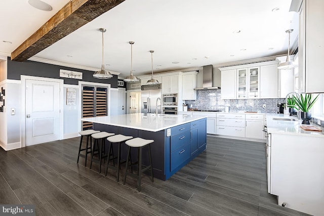 kitchen with white cabinets, decorative light fixtures, a kitchen island with sink, wall chimney exhaust hood, and appliances with stainless steel finishes