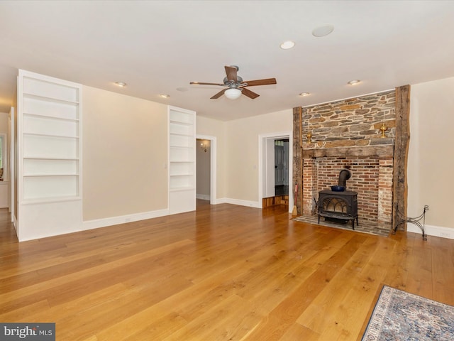 unfurnished living room with built in shelves, ceiling fan, a wood stove, and light wood-type flooring