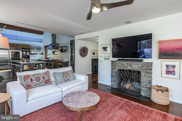 living room with a stone fireplace, dark hardwood / wood-style floors, ceiling fan, and sink