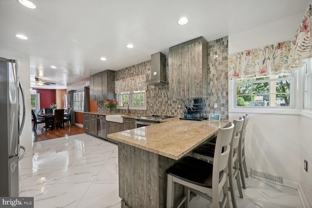 kitchen featuring kitchen peninsula, backsplash, ceiling fan, wall chimney range hood, and sink