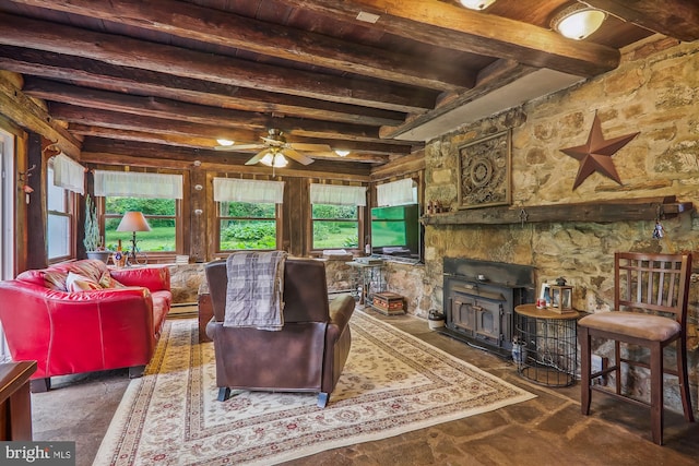 living room featuring beamed ceiling, ceiling fan, a stone fireplace, and a wood stove