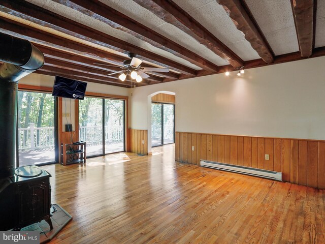 unfurnished living room featuring ceiling fan, baseboard heating, light hardwood / wood-style floors, beamed ceiling, and a wood stove