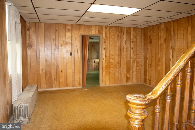 unfurnished room with radiator, wooden walls, a drop ceiling, and carpet