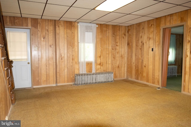 unfurnished room featuring carpet flooring, a drop ceiling, radiator, and wood walls