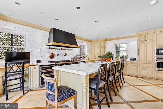 kitchen with appliances with stainless steel finishes, backsplash, light tile flooring, wall chimney range hood, and a kitchen island with sink