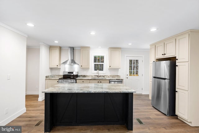 kitchen with light stone counters, cream cabinets, wall chimney exhaust hood, light wood-type flooring, and stainless steel appliances