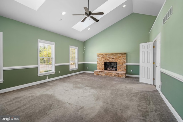 unfurnished living room featuring a skylight, ceiling fan, a stone fireplace, high vaulted ceiling, and dark colored carpet