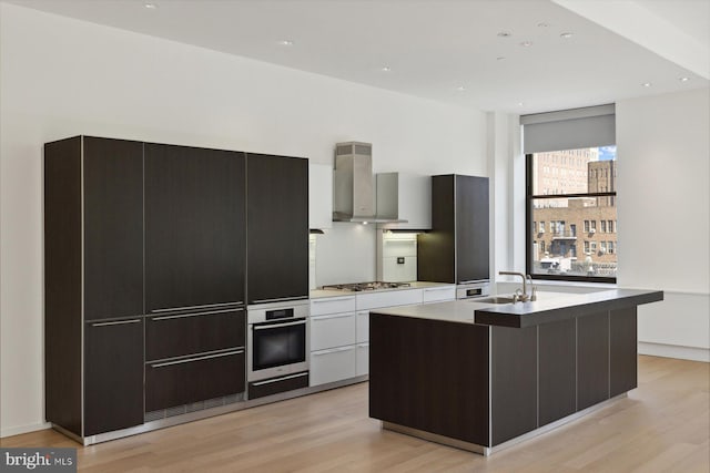 kitchen featuring white cabinetry, appliances with stainless steel finishes, wall chimney range hood, an island with sink, and modern cabinets