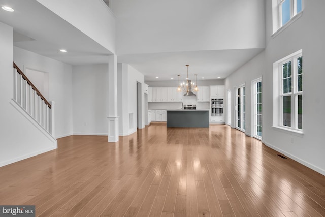 unfurnished living room with a notable chandelier, light wood finished floors, a high ceiling, baseboards, and stairs