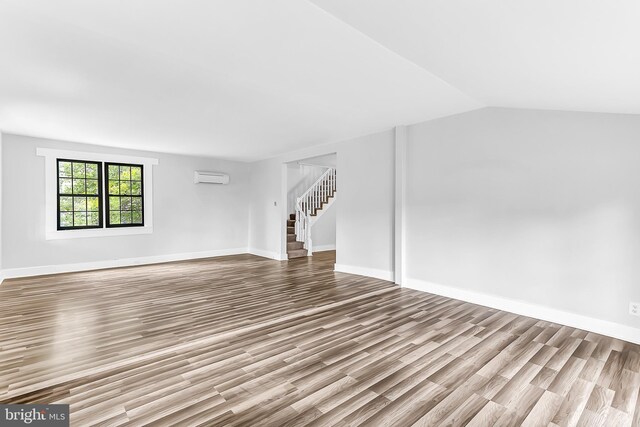 unfurnished living room with light wood-style flooring, a wall mounted AC, vaulted ceiling, baseboards, and stairs