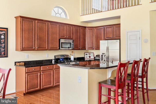 kitchen with appliances with stainless steel finishes, light hardwood / wood-style floors, a high ceiling, dark stone counters, and a breakfast bar