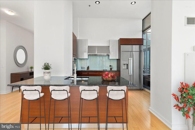 kitchen with wall chimney exhaust hood, light hardwood / wood-style flooring, stainless steel appliances, tasteful backsplash, and dark stone counters