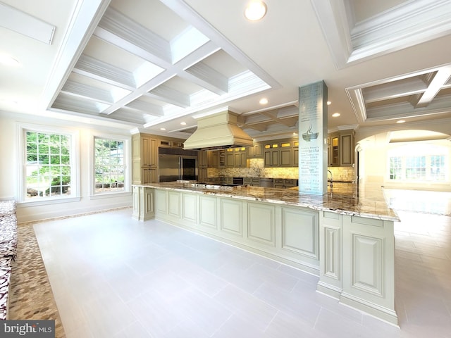 kitchen with light stone countertops, backsplash, custom range hood, a large island with sink, and built in fridge