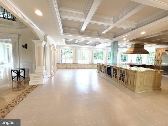 kitchen with coffered ceiling, a spacious island, light stone counters, custom range hood, and decorative columns