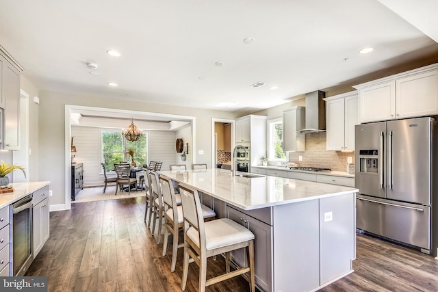 kitchen with backsplash, stainless steel appliances, wall chimney range hood, an island with sink, and dark hardwood / wood-style flooring