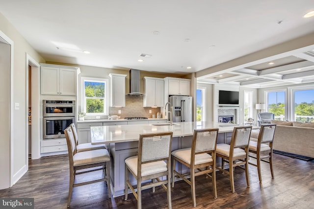 kitchen featuring stainless steel appliances, a center island with sink, wall chimney range hood, coffered ceiling, and dark hardwood / wood-style flooring