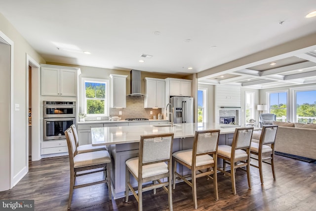 kitchen featuring stainless steel appliances, dark wood-type flooring, wall chimney range hood, coffered ceiling, and a fireplace