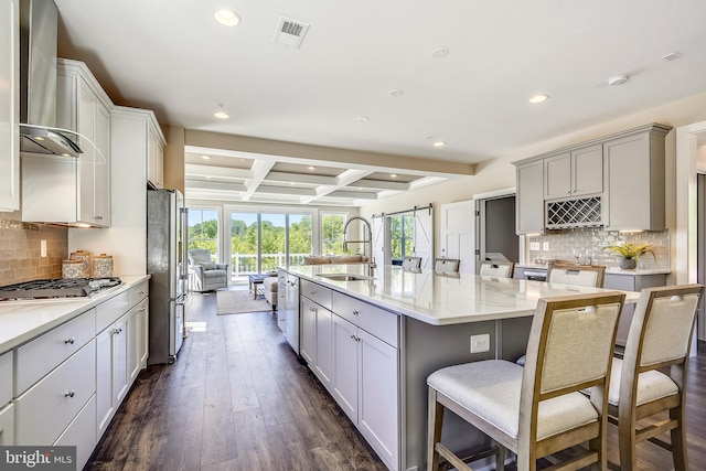 kitchen with wall chimney exhaust hood, dark hardwood / wood-style floors, beam ceiling, a center island with sink, and coffered ceiling