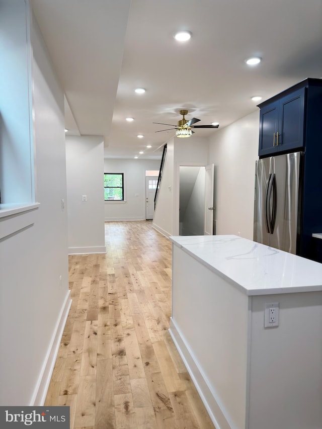 kitchen with stainless steel fridge, light stone counters, ceiling fan, blue cabinetry, and light hardwood / wood-style flooring