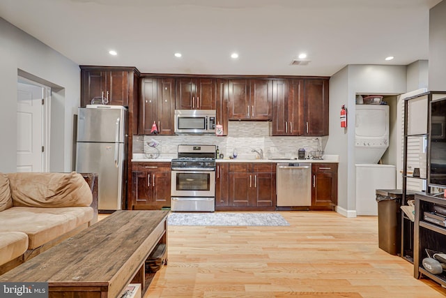 kitchen featuring dark brown cabinets, stacked washer / dryer, appliances with stainless steel finishes, light hardwood / wood-style floors, and tasteful backsplash