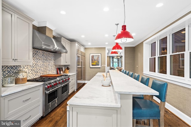 kitchen with pendant lighting, wall chimney range hood, light stone countertops, premium appliances, and a kitchen bar