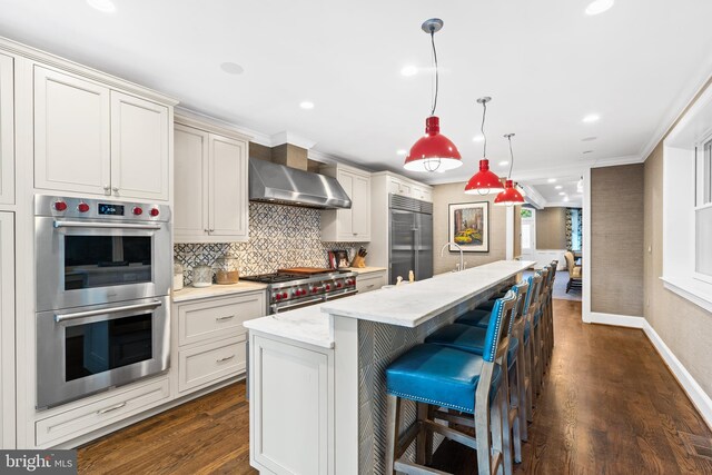 kitchen featuring dark wood-type flooring, wall chimney range hood, premium appliances, decorative light fixtures, and a center island with sink