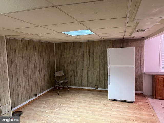 finished basement featuring a paneled ceiling, light wood finished floors, visible vents, and freestanding refrigerator