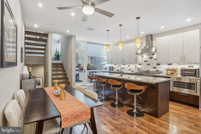 kitchen featuring light wood-type flooring, a center island with sink, hanging light fixtures, and wall chimney range hood