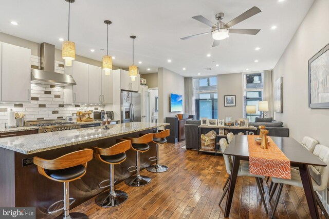 kitchen with a breakfast bar, wall chimney range hood, dark hardwood / wood-style floors, appliances with stainless steel finishes, and decorative light fixtures