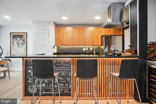 kitchen featuring wall chimney range hood, stainless steel fridge, light wood-type flooring, a breakfast bar, and tasteful backsplash