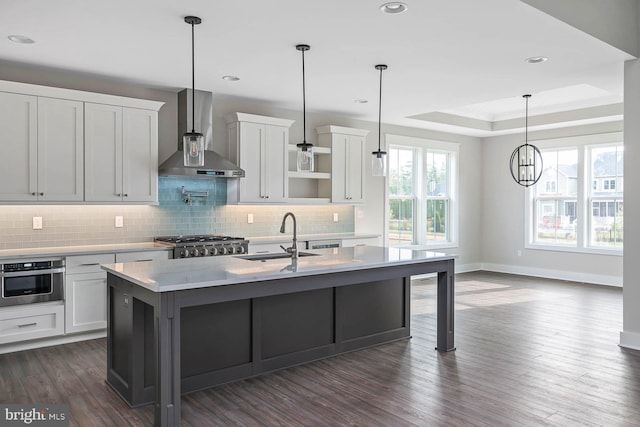 kitchen with an island with sink, wall chimney exhaust hood, stainless steel oven, dark wood-type flooring, and sink