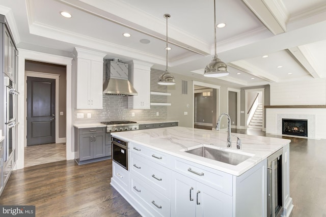 kitchen with sink, white cabinetry, hanging light fixtures, an island with sink, and wall chimney exhaust hood