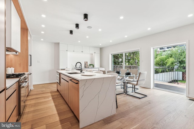 kitchen with high end stainless steel range oven, sink, a center island with sink, light hardwood / wood-style flooring, and white cabinetry