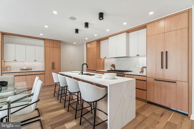 kitchen with light wood-type flooring, tasteful backsplash, sink, a center island with sink, and white cabinets