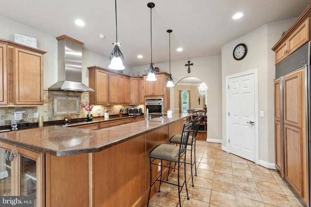 kitchen with wall chimney range hood, light tile flooring, backsplash, a center island with sink, and paneled built in refrigerator